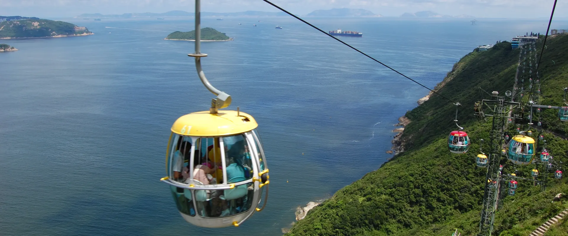 A yellow cable car cabin with passengers hanging from its cable above the sea, with a green headland on the right